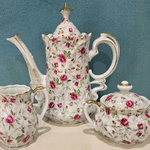 May include: A porcelain tea set featuring a floral pattern of red roses and green leaves on a white background. The set includes a teapot, creamer, and sugar bowl, all with gold trim.