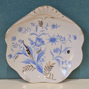 May include: White porcelain decorative plate with a scalloped edge and gold trim. The plate features a blue floral design with gold and brown accents. The flowers and leaves are painted in a delicate style, creating an elegant aesthetic.