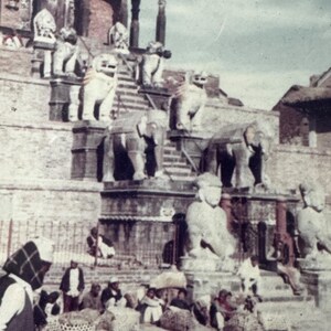 May include: A stone temple with a staircase leading up to the entrance. There are several statues of elephants and lions on the steps. People are gathered at the bottom of the stairs.