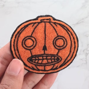 May include: Orange felt pumpkin patch appliqué with a black stitched outline and a scary face. The pumpkin has two large eyes, a wide mouth with teeth, and a stem on top.