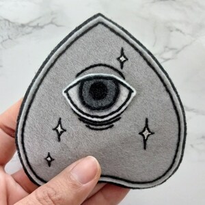 May include: A gray felt patch in the shape of a heart with an embroidered black eye and white stars.