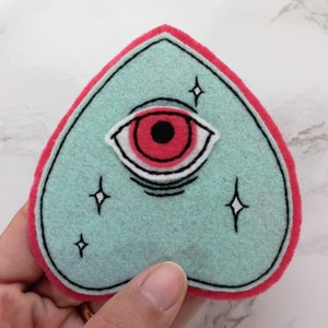 May include: A teal felt patch in the shape of a heart with a red eye and white eyelashes. The eye has a black pupil and is surrounded by white stars.