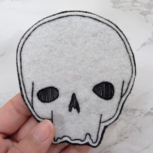 May include: A white felt skull patch with black embroidered details. The skull has two eye sockets, a nose, and a mouth.