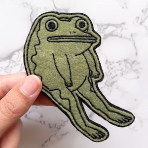May include: A green felt frog with black outlines, sitting with its legs outstretched. The frog has a large, round head with big eyes and a wide mouth.