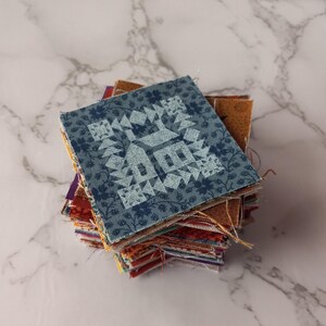 May include: A stack of square fabric quilt blocks featuring a variety of patterns and colors. The top block is a blue fabric with a light blue printed design of a house surrounded by geometric shapes and floral motifs, with frayed edges.