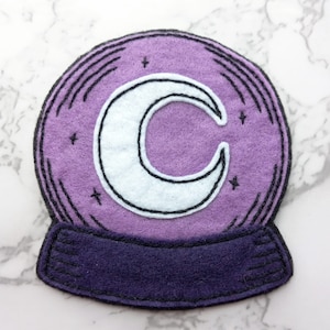 May include: Purple felt snow globe with a white crescent moon and black stitching. The snow globe has a dark purple felt base.