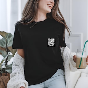 May include: A black t-shirt with a white QR code printed on the left chest pocket. The QR code is surrounded by a white border.