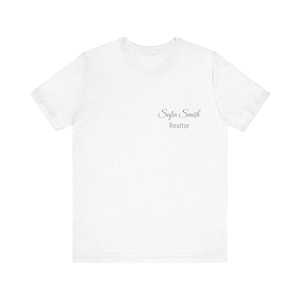 Puede incluir: Camiseta blanca con el texto "Sofia Smith Realtor" impreso en una escritura gris oscuro en la parte superior izquierda del pecho. La camiseta tiene mangas cortas y cuello redondo.