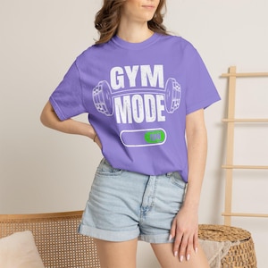 Könnte beinhalten: Ein lila T-Shirt mit dem weißen Schriftzug "GYM MODE" und einer Hantel-Grafik. Darunter befindet sich ein horizontaler Balken mit einer grünen "ON"-Anzeige. Das Shirt wird mit Jeansshorts getragen.