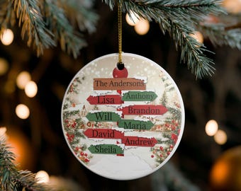 May include: A circular Christmas tree decoration featuring a winter landscape and the family name "The Andersons." The ornament has directional signs with family member names. It hangs from a gold string and has a red bulb.