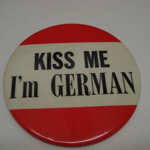 May include: A red and white button with the text "Kiss Me I'm German" in black.