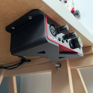May include: Black 3D printed bracket mounted under a desk, holding a red Scarlett 2i2 audio interface with knobs and ports.