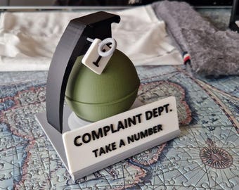 May include: A green and black 3D printed model of a grenade with a white sign that reads "Complaint Dept. Take a Number".