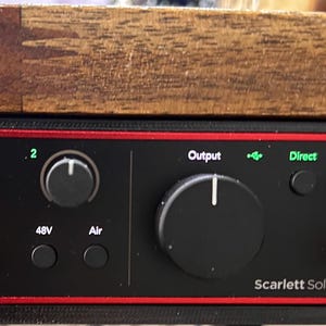 May include: Black Scarlett Solo audio interface with red trim, featuring multiple knobs and input jacks. The interface has labeled controls for input levels, output, and direct monitoring. A headphone jack is visible.