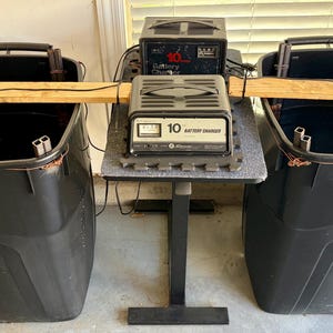 May include: Two large black plastic bins connected by a wooden board, each containing metal rods. Between the bins, two black battery chargers are stacked on a small stand. The front charger features a gauge and the text 10 BATTERY CHARGER.