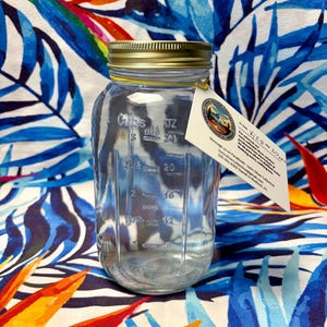 May include: A clear glass jar with a gold-colored metal lid and embossed measurement markings in cups and ounces. A white paper tag is attached to the neck, featuring the Ionic Silver Collective logo and text details about the solution.