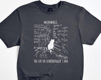 May include: Dark gray t-shirt with a white graphic of a cat and mathematical equations. The text reads "MEANWHILE, THE CAT IN SCHRÖDINGER'S BOX". The shirt is a crew neck style.