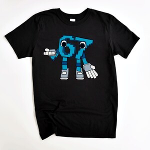 May include: Black t-shirt featuring a pixelated cartoon character in shades of blue, gray, and white. The character is a stylized representation of the numbers "67" with arms, legs, and gloves. The design is centered on the shirt.