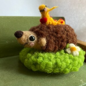 May include: A brown hedgehog with a yellow and red caterpillar on its back, sitting on a green felt cushion with a white flower.