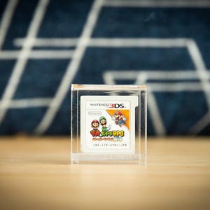 May include: A Nintendo 3DS game cartridge, featuring the title "Mario & Luigi: Paper Jam Bros." in a clear acrylic display case. The game's label shows Mario, Luigi, and other characters. The background is a blurred blue and white pattern.