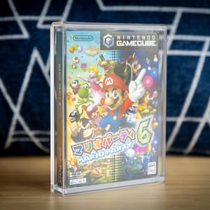 May include: A Nintendo GameCube game case for Mario Party 6. The game case features a colorful illustration of Mario and other characters. The case is in a clear protective case, sitting on a wooden surface.