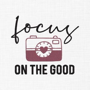 May include: A white background with the words "focus" and "ON THE GOOD" in black script. A maroon camera illustration with a heart-shaped lens is centered between the words. The image has a textured appearance.
