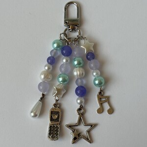May include: A beaded keychain with a silver clasp. The keychain features strands of white, lavender, and teal beads, with silver star and music note charms, and a small flip phone charm.