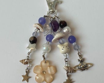 May include: A silver keychain with various charms. The charms include a flower, stars, seashells, a planet, and beads in shades of purple, white, and brown. The keychain is on a white background.