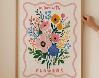 Op de afbeelding: Een ingelijst print met een roze rand en een witte achtergrond met een boeket kleurrijke bloemen met de tekst "in love with flowers" in roze.