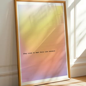 Könnte beinhalten: Ein gerahmter Kunstdruck mit einem Farbverlauf aus Gelb-, Orange- und Rosatönen. Der Holzrahmen umgibt das Kunstwerk, das den Text "Stay close to what feels like sunshine" in einer einfachen Schriftart enthält. Der Druck lehnt an einer weißen Wand.