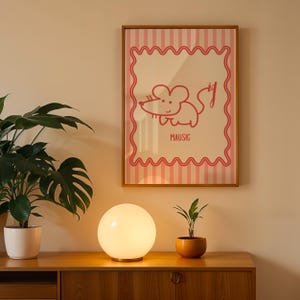 May include: A framed poster with a pink and white striped design and a red cartoon mouse illustration. The word "MAUSIG" is below the illustration. A white globe lamp and potted plants sit on a wooden cabinet.