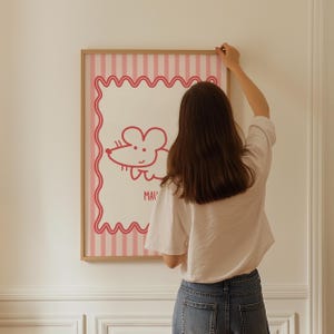 May include: A framed art print with a cartoon mouse illustration in red on a white background. The print has a pink wavy border and a pink striped background. The frame is made of light brown wood.