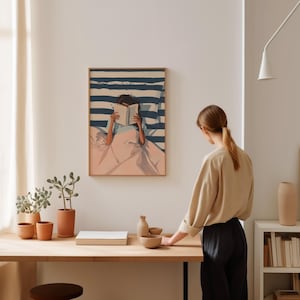 A woman is standing in a room, looking at a framed painting of a person reading a book. The painting is hanging on the wall above a wooden table with various items on it, including a book, a vase, and a bowl.