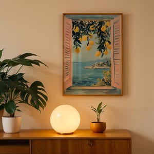 a wooden cabinet with a round table lamp on top, a framed painting of a window overlooking a beach, and two potted plants on the cabinet.