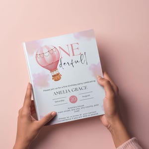 Può includere: Un invito di compleanno rosa e bianco con un'illustrazione di mongolfiera. L'invito recita "ONE derful!" e include i dettagli per la festa del primo compleanno di Amelia Grace, inclusi data e ora.