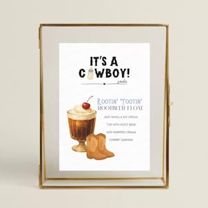 May include: A framed print with the text "It's a Cowboy!" and a recipe for a Rootin' Tootin' Rootbeer Float. The recipe includes vanilla ice cream, root beer, whipped cream, and a cherry garnish. A pair of cowboy boots is also shown.