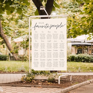 May include: A white sign with black text that reads "Our favorite people" and lists table numbers from one to twenty. The sign is hanging from a metal stand in a garden setting.