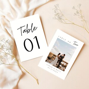 May include: Two white table number cards with black calligraphy. The first card says "Table 01". The second card says "Table No. Two" and features a photo of a wedding couple.