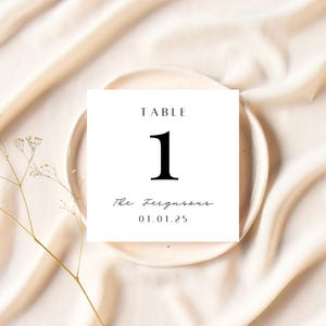 May include: A white table number card with the word "TABLE" at the top and the number "1" in a bold font. Below, the card reads "The Fergusons" and the date "01.01.25". The card rests on a beige plate, set against a cream-colored fabric.
