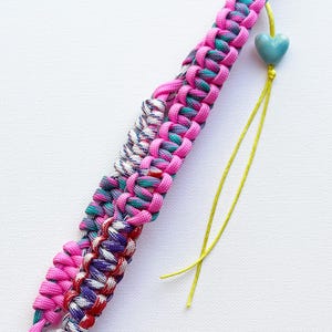 May include: A braided paracord bracelet featuring a vibrant pink cord interwoven with a multi-colored cord in shades of purple, teal, and red. A single light blue heart-shaped bead hangs from a thin yellow string attached to the main braid.