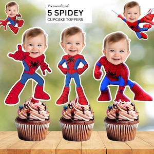 May include: Five Spidey cupcake toppers with a baby's face, in red, blue, and black. The cupcakes have chocolate frosting, sprinkles, and chocolate drizzle. The text reads "Personalized 5 SPIDEY CUPCAKE TOPPERS."