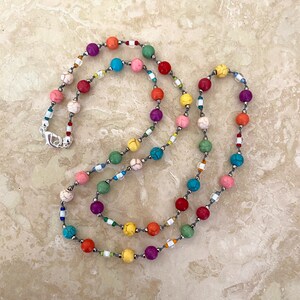 May include: A colorful beaded necklace with a variety of colors including pink, red, orange, yellow, green, blue, and purple. The beads are strung on a gray cord with silver accents.