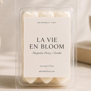 Pu&ograve; includere: Una confezione in plastica trasparente contenente cialde di cera a forma di cuore. L'etichetta riporta Adorably Tiny, La Vie En Bloom, Magnolia Peony + Vanilla, soy wax | 70,8g e adorablytiny.com. La cera &egrave; bianco sporco e disposta a griglia.