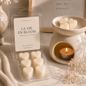 Pu&ograve; includere: Una confezione in plastica trasparente con sei cialde di cera di soia a forma di cuore color crema, etichettata LA VIE EN BLOOM Magnolia Peony Vanilla 2.7oz, accanto a un bruciatore in ceramica maculata con tre cuori e una candela accesa.