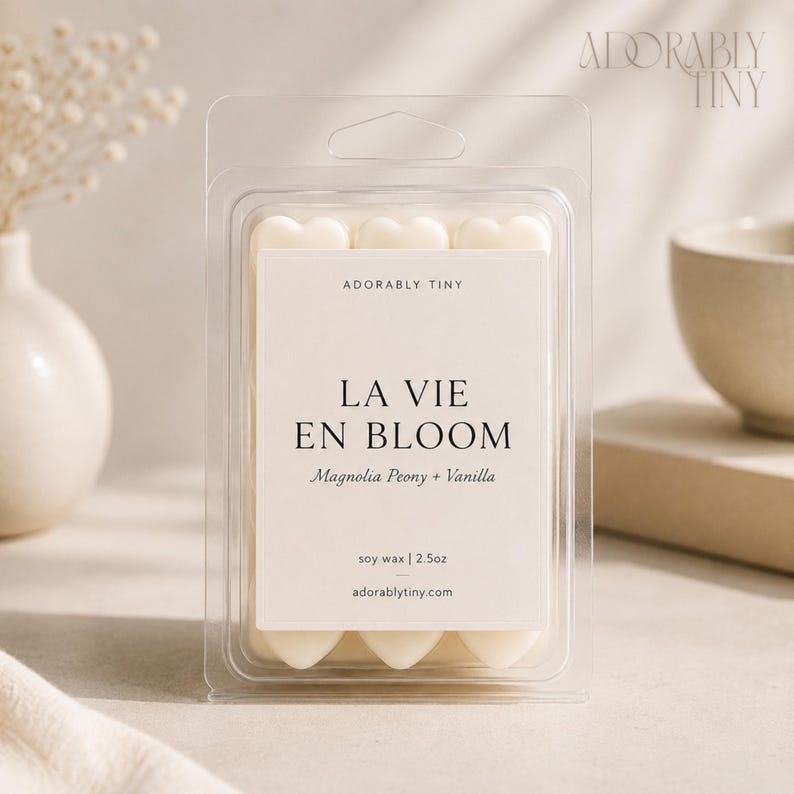 Pu&ograve; includere: Una confezione in plastica trasparente contenente cialde di cera a forma di cuore. L'etichetta riporta Adorably Tiny, La Vie En Bloom, Magnolia Peony + Vanilla, soy wax | 70,8g e adorablytiny.com. La cera &egrave; bianco sporco e disposta a griglia.