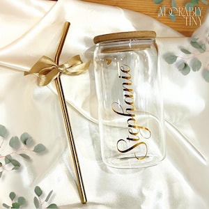 May include: A clear glass can-shaped drinking glass with a bamboo lid and the name "Stephanie" in gold script. A gold-colored metal straw with a gold bow is next to the glass. The background is white fabric with greenery.
