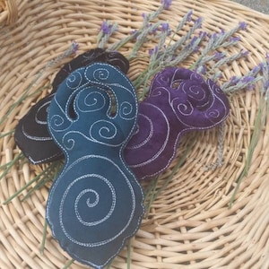 May include: Three decorative, abstract-shaped fabric items in black, teal, and purple, each with white embroidered spiral designs. They rest on a woven basket with sprigs of lavender. The basket is made of light brown wicker.
