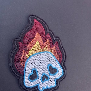 May include: Embroidered patch of a white skull with heart-shaped eyes on fire. The flames are red, orange, and yellow.