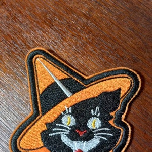 May include: A black cat wearing a witch's hat and a bow tie. The cat is embroidered on a patch with orange and black fabric.