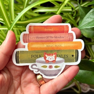 May include: A sticker featuring a stack of books with titles like "Poems" and "Folk Tales." A teacup with a fox peeking out is at the bottom. The sticker has a whimsical, storybook aesthetic, with warm colors and detailed illustrations.